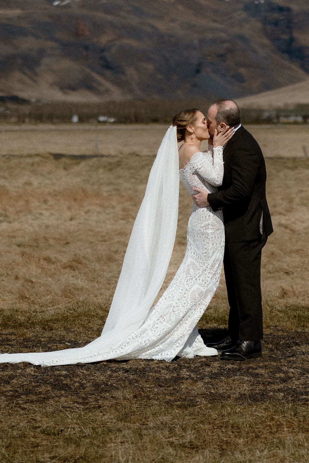 Bride and groom sharing a kiss outdoors, wearing elegant wedding attire.