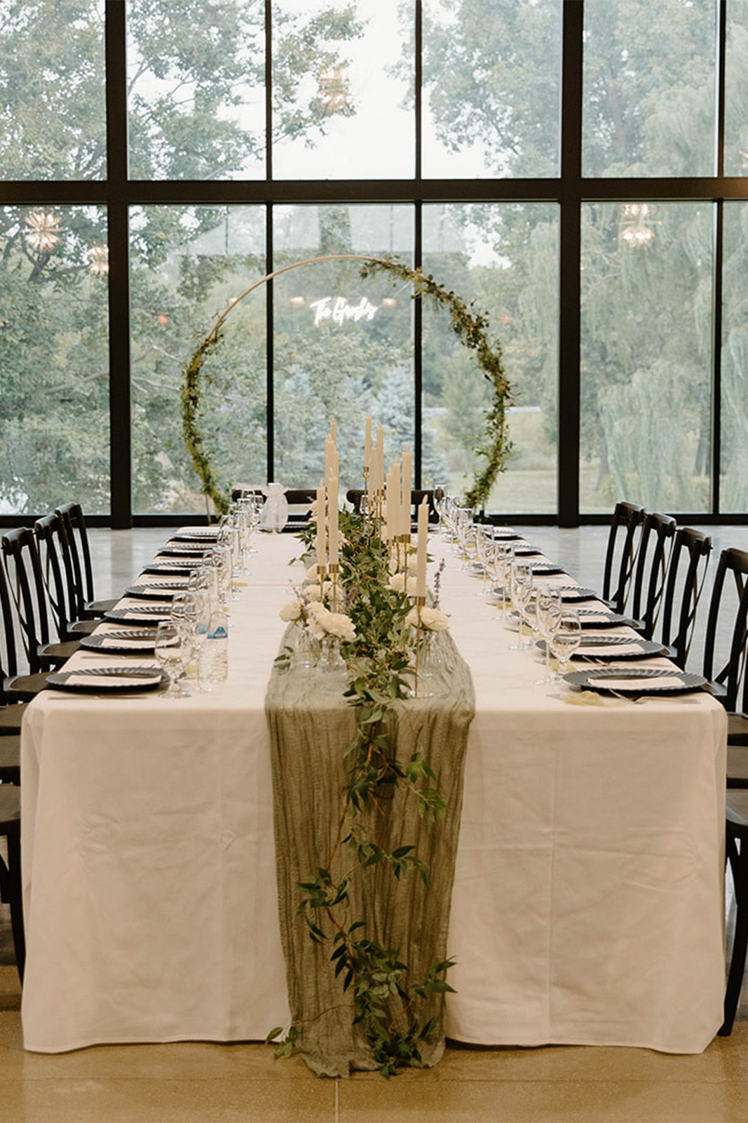 Elegant dining table set with greenery and candles near large windows.