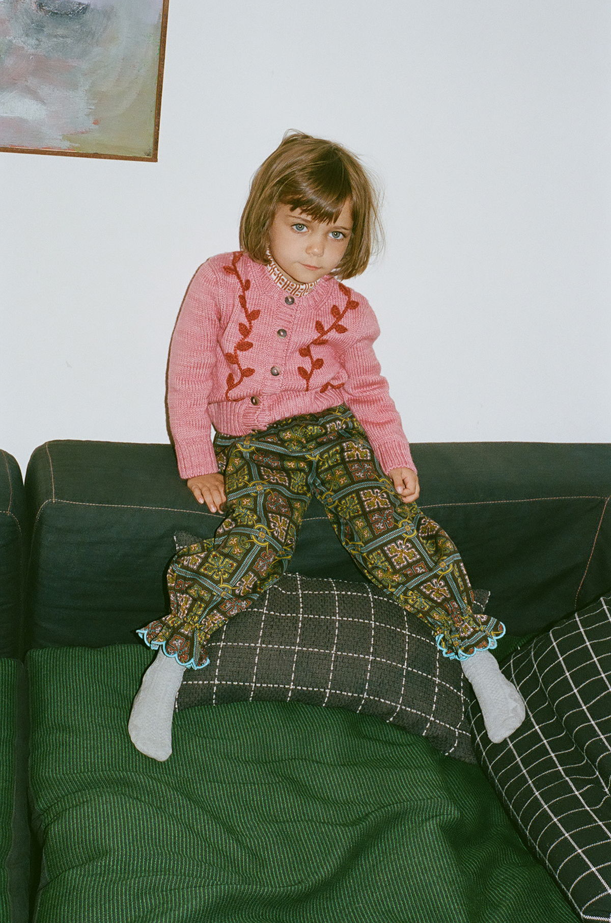 Child sitting on a couch, wearing a pink sweater and patterned pants.
