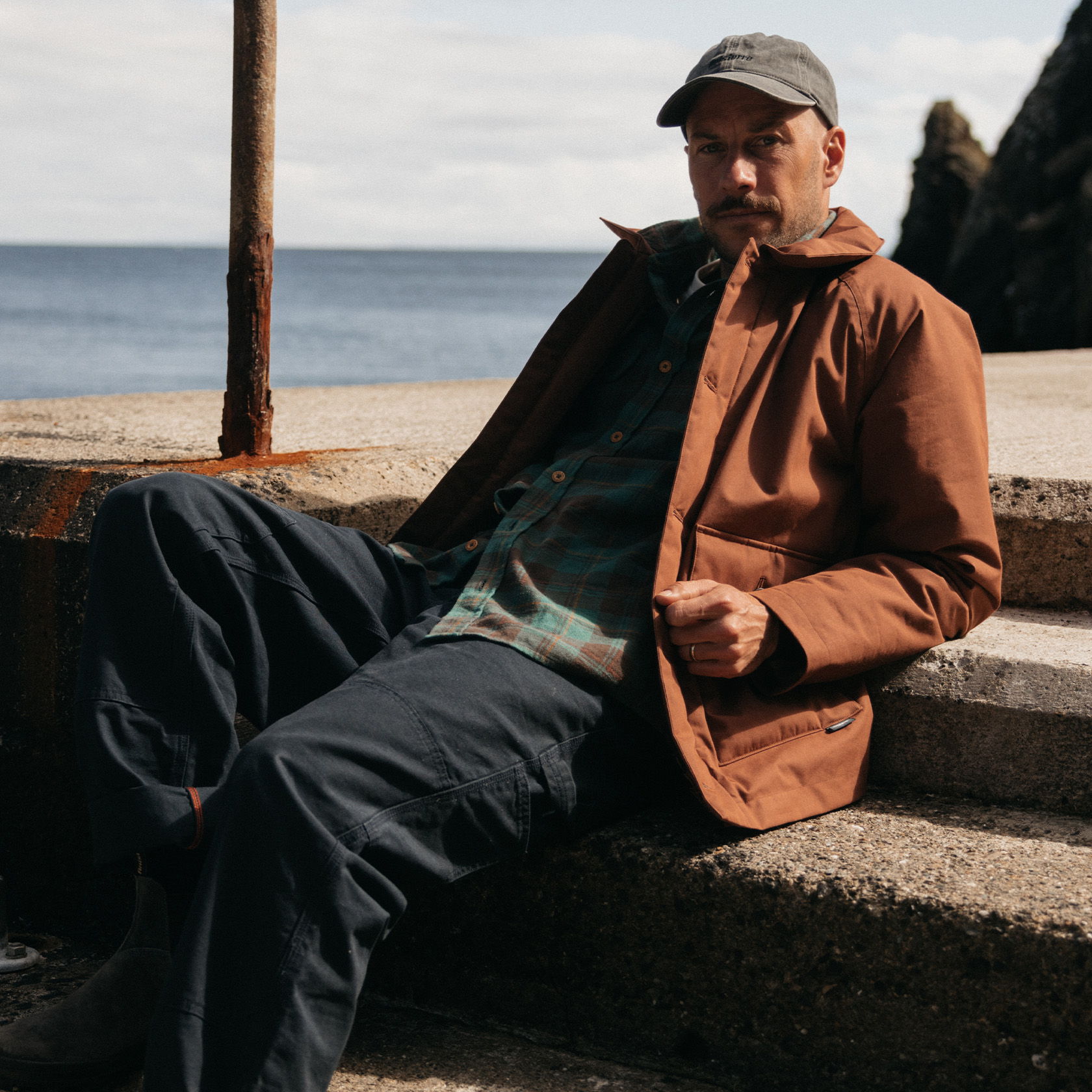 Man sitting on steps by the water, wearing a brown jacket and cap.