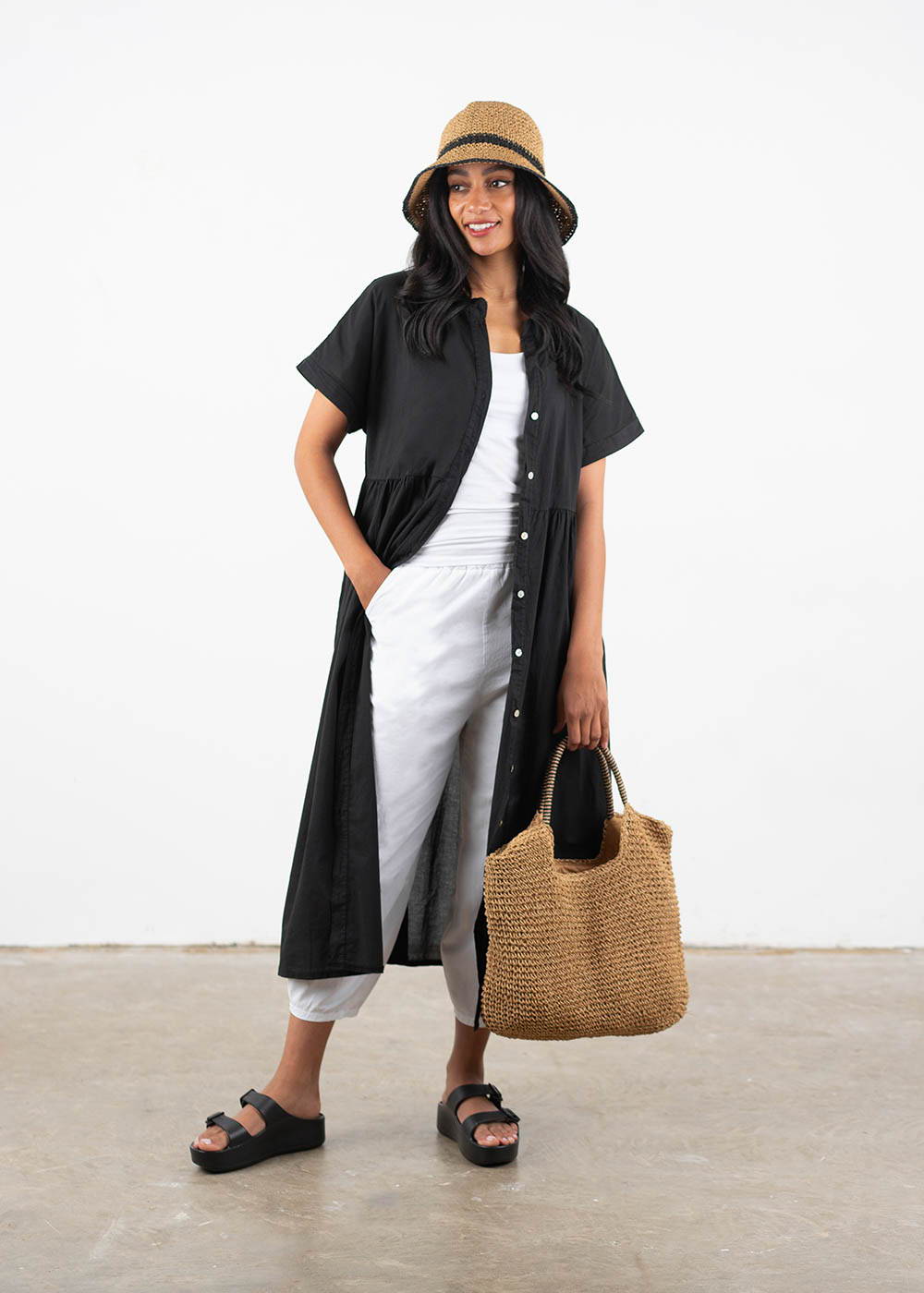 A model wearing a pair of white barrell trousers with tapered leg, black shortsleeved long shirt dress, white v neck top, black chunky platform slides, a straw hat and bag