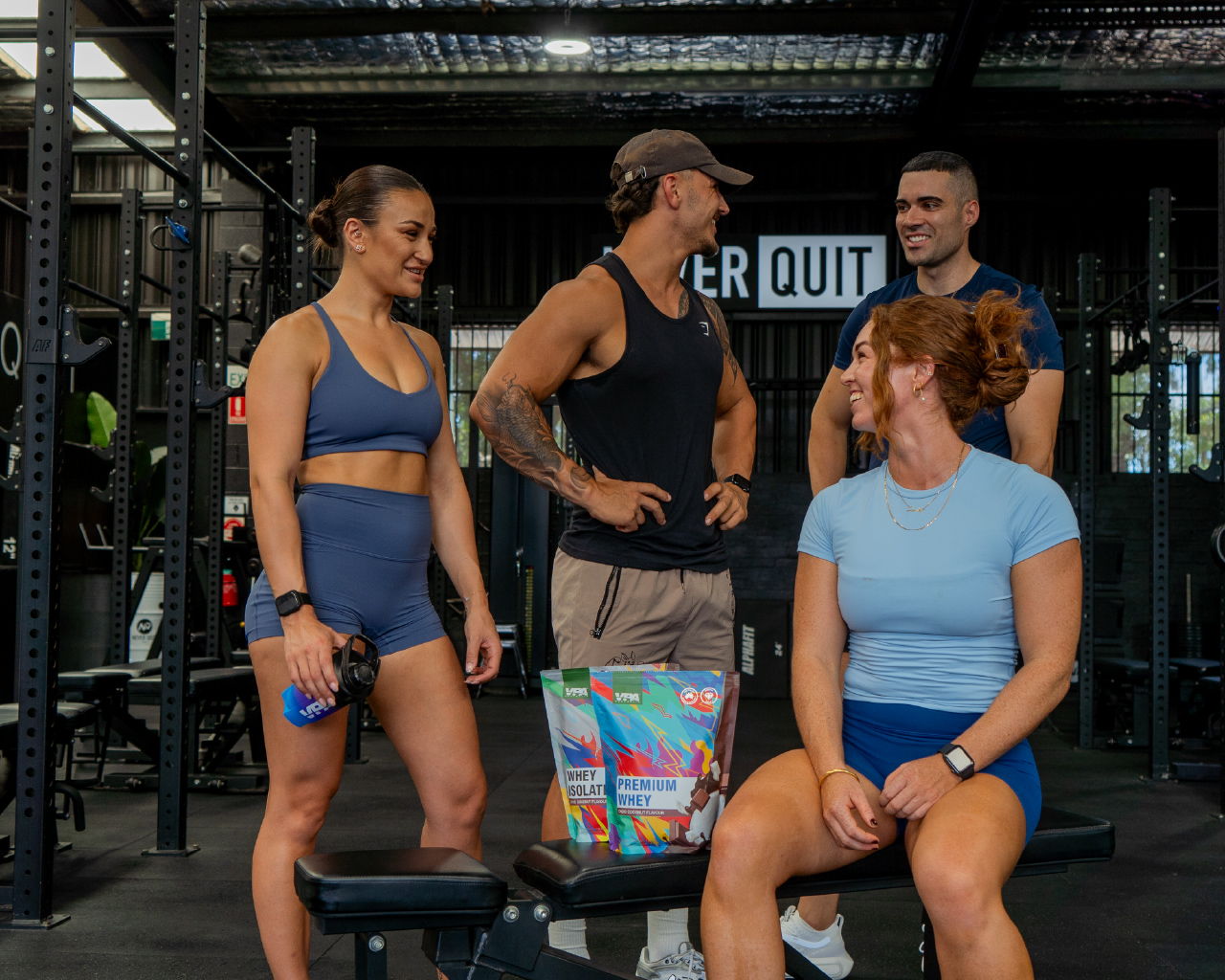 Four people chatting in a gym, two sitting and two standing nearby.