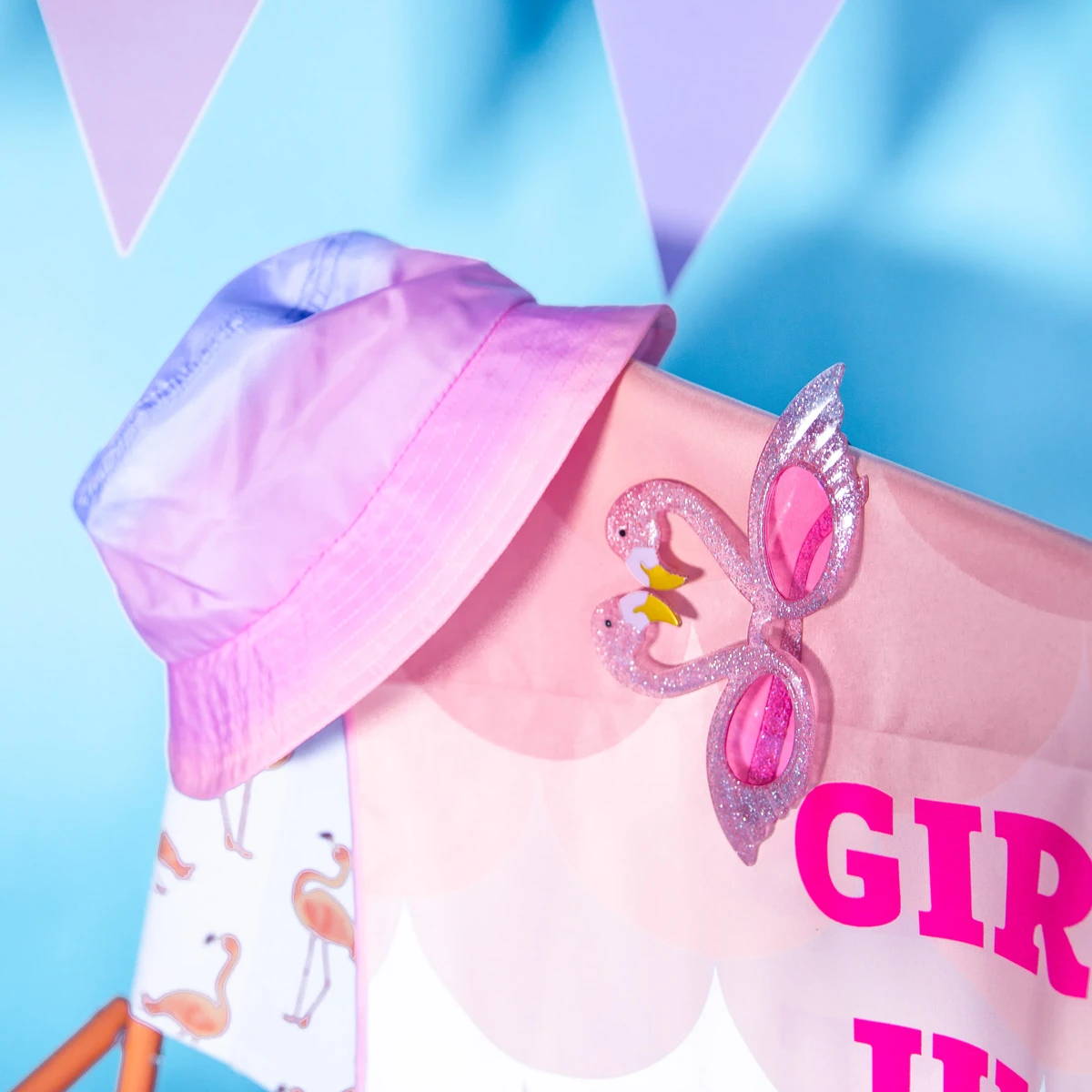 Flamingo-themed party accessories including a pink bucket hat and glittery flamingo glasses on a draped pink fabric