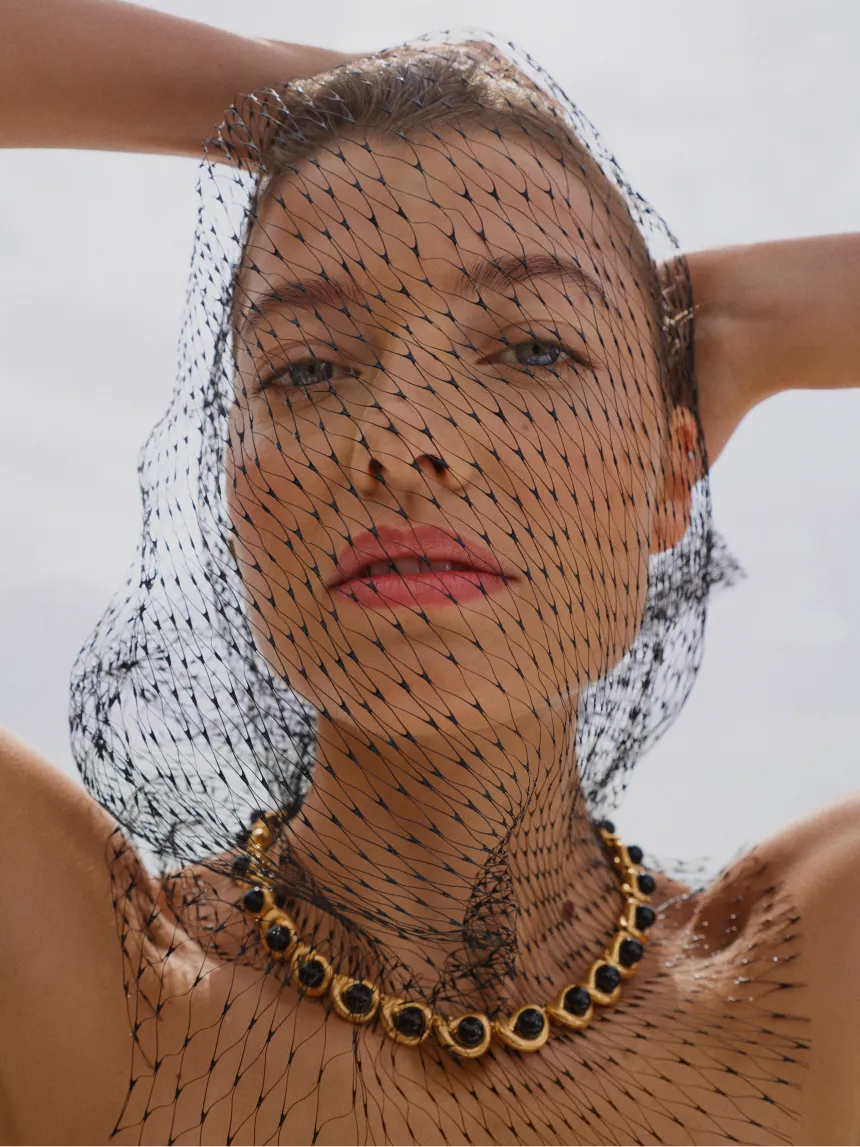 Woman with a black net veil and gold necklace against a light background.