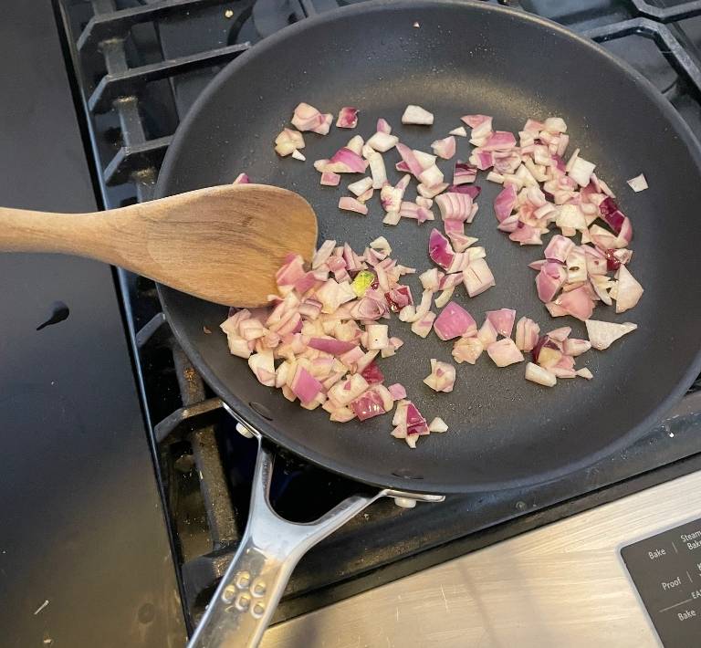 An unseen hand uses a wooden spoon to stir chopped red onion in a Misen Nonstick Pan, which sits on a stovetop.