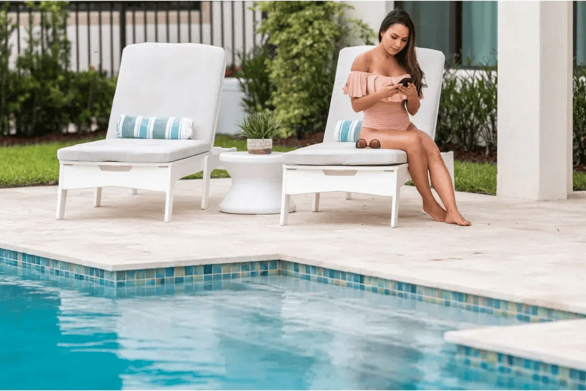 Two white poolside loungers with a round side table on a light stone patio beside a swimming pool, with landscaping behind.
