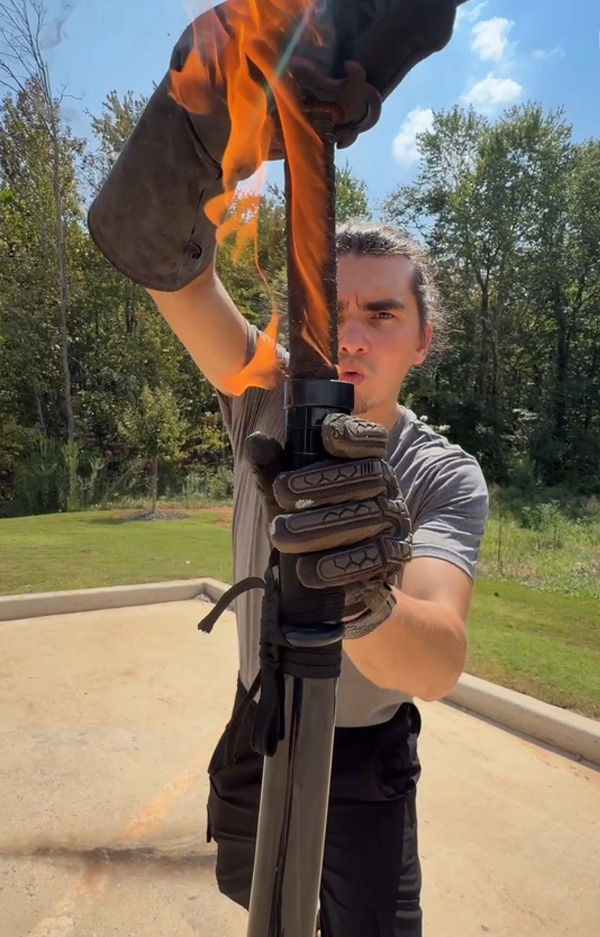 Person holding a flaming object outdoors, wearing gloves and a focused expression.