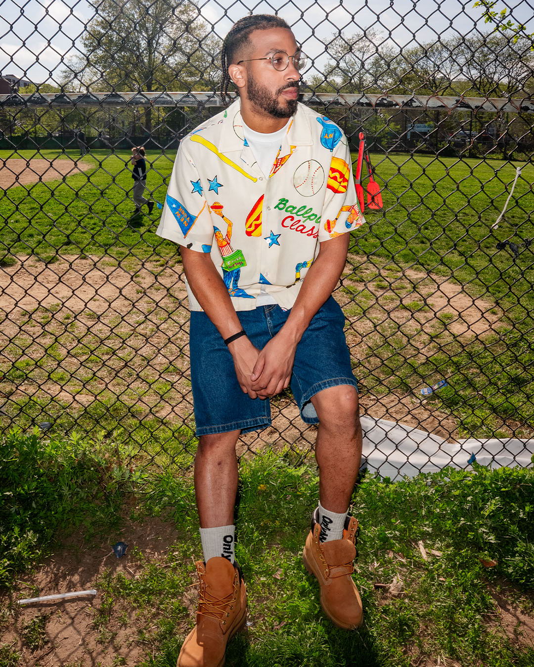 Person in colorful shirt and shorts, leaning against a fence near a field.