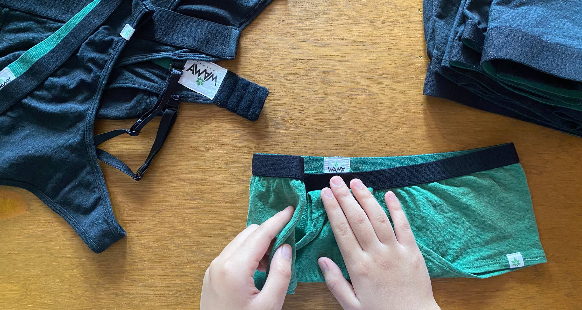 two hands folding a green pair of underwear on a wooden table with stacks of black underwear and bras nearby