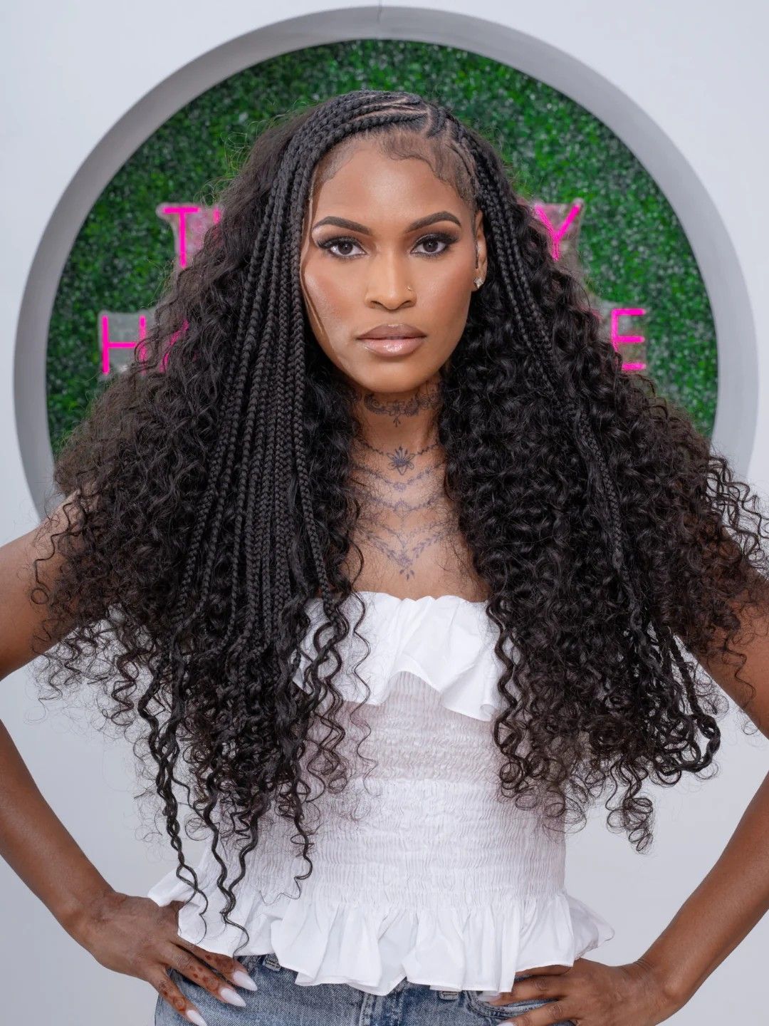 A woman with long, curly hair extensions wearing a white top, posing confidently.