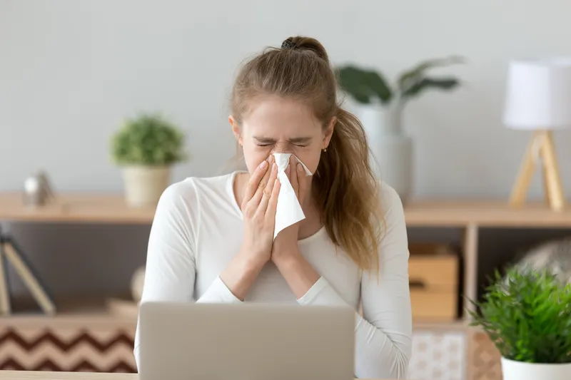 Een jonge vrouw die verkouden is of last heeft van een allergie niest in een tissue terwijl ze achter een laptop aan haar bureau of eettafel zit. Allergie kan heel belastend zijn tijdens je werk en schoolactiviteiten.