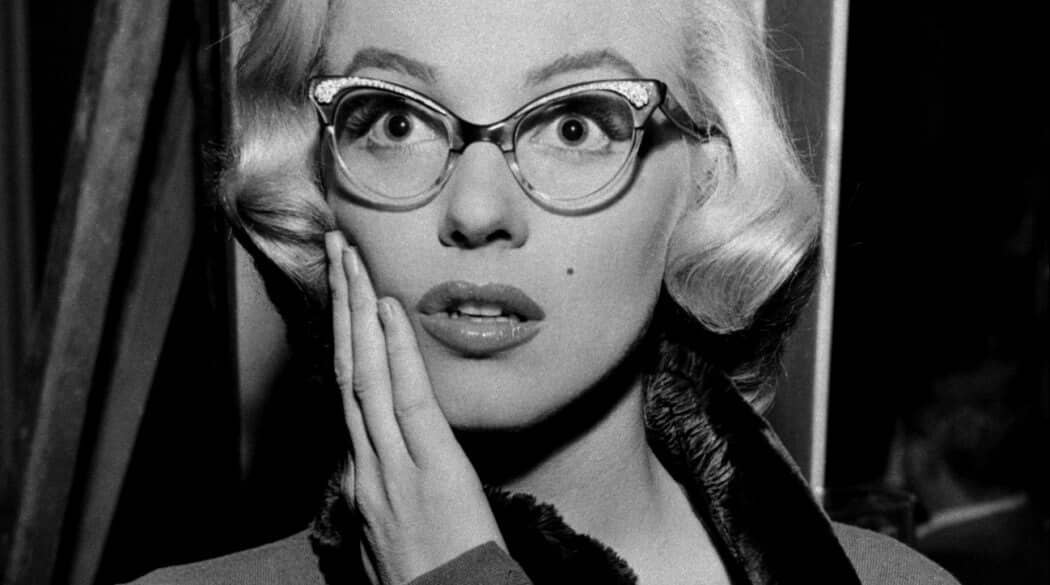 A black and white photo of Icon Marilyn Monroe wearing cat-eye glasses while putting her hand on her cheek