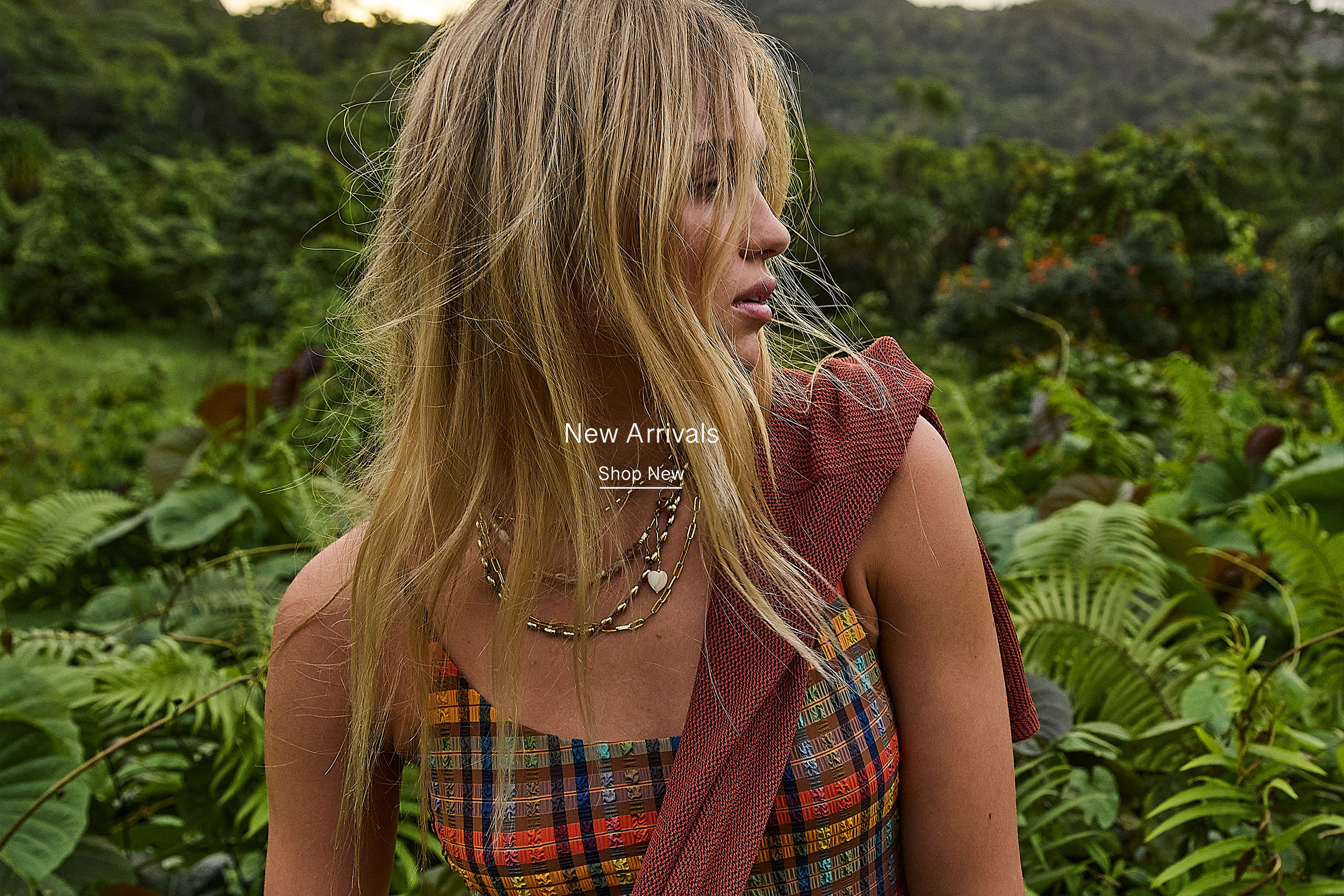 A woman with long hair in a plaid outfit, surrounded by greenery.