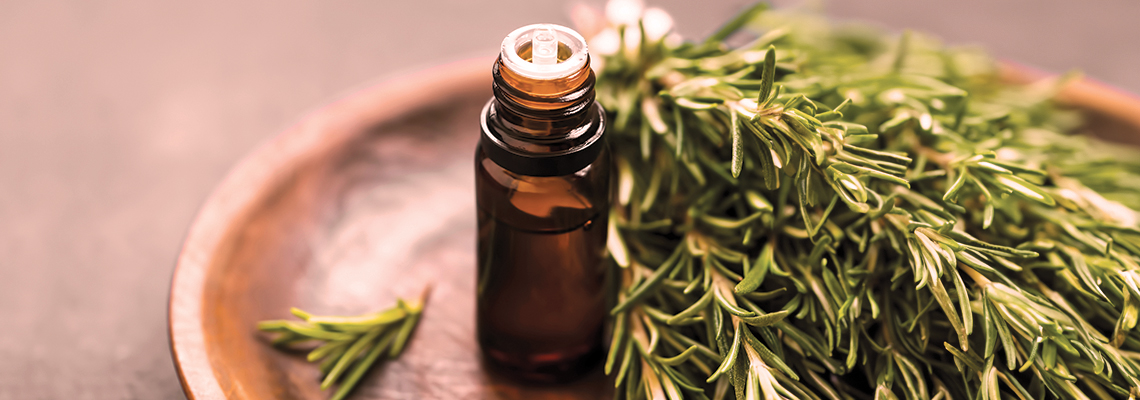 Brown essential oil bottle next to fresh rosemary sprigs on a wooden plate.