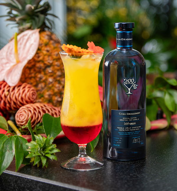 Colorful cocktail beside a decorative bottle with tropical fruit in the background.