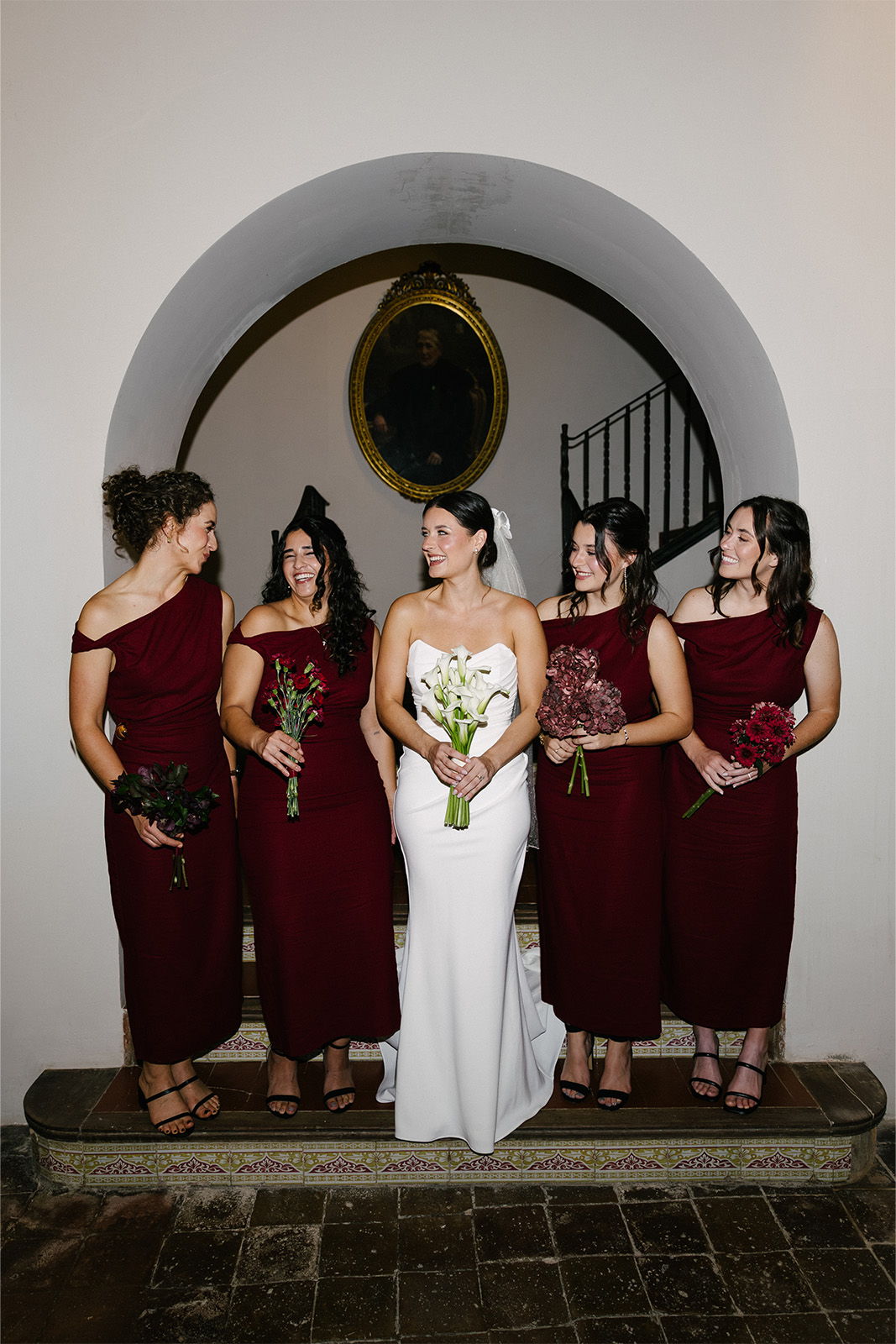 Bride and bridesmaids in matching dresses, smiling in an elegant indoor setting.