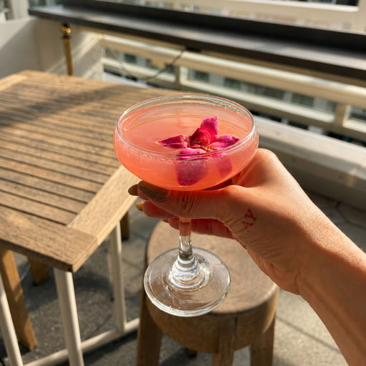 A hand holding a pink cocktail with flower garnish on a sunny balcony.