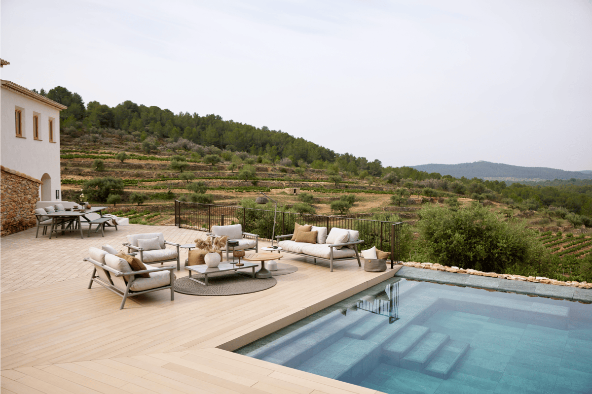 Modern outdoor lounge and dining furniture on a poolside terrace overlooking rolling hills, featuring neutral cushions, round rugs, and a relaxed resort-style layout.