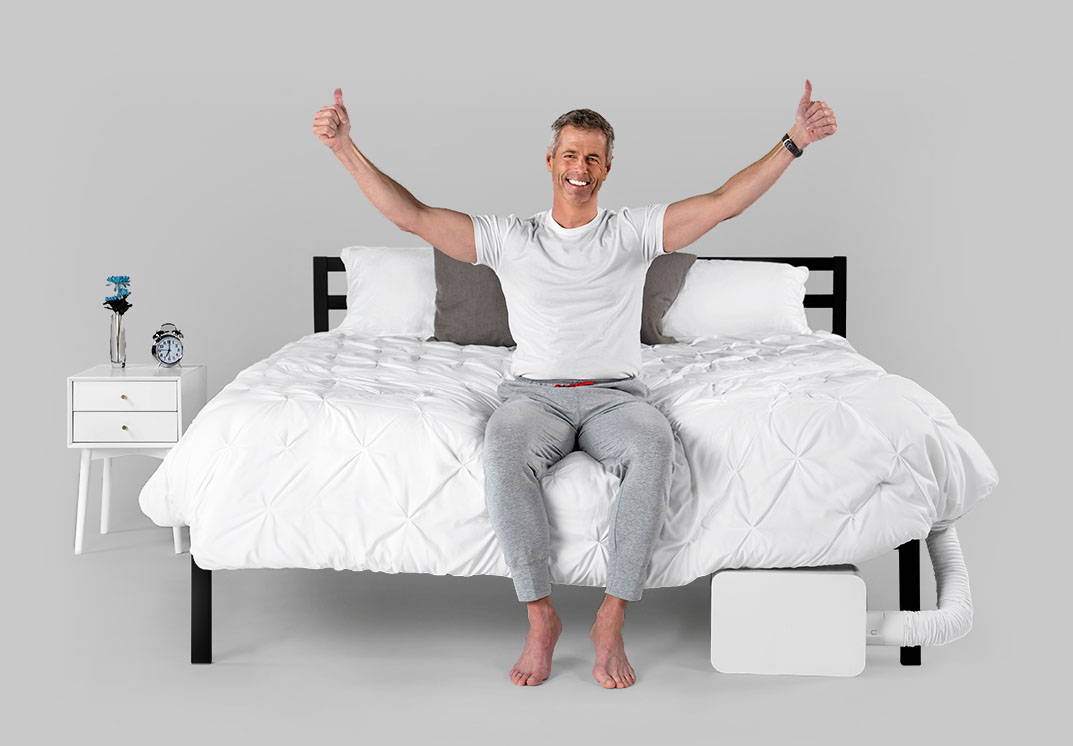 An older man in a white tshirt and gray sweatpants sitting on a bed with his hands raised up and giving thumbs up, and a BedJet is attached to the foot of the bed