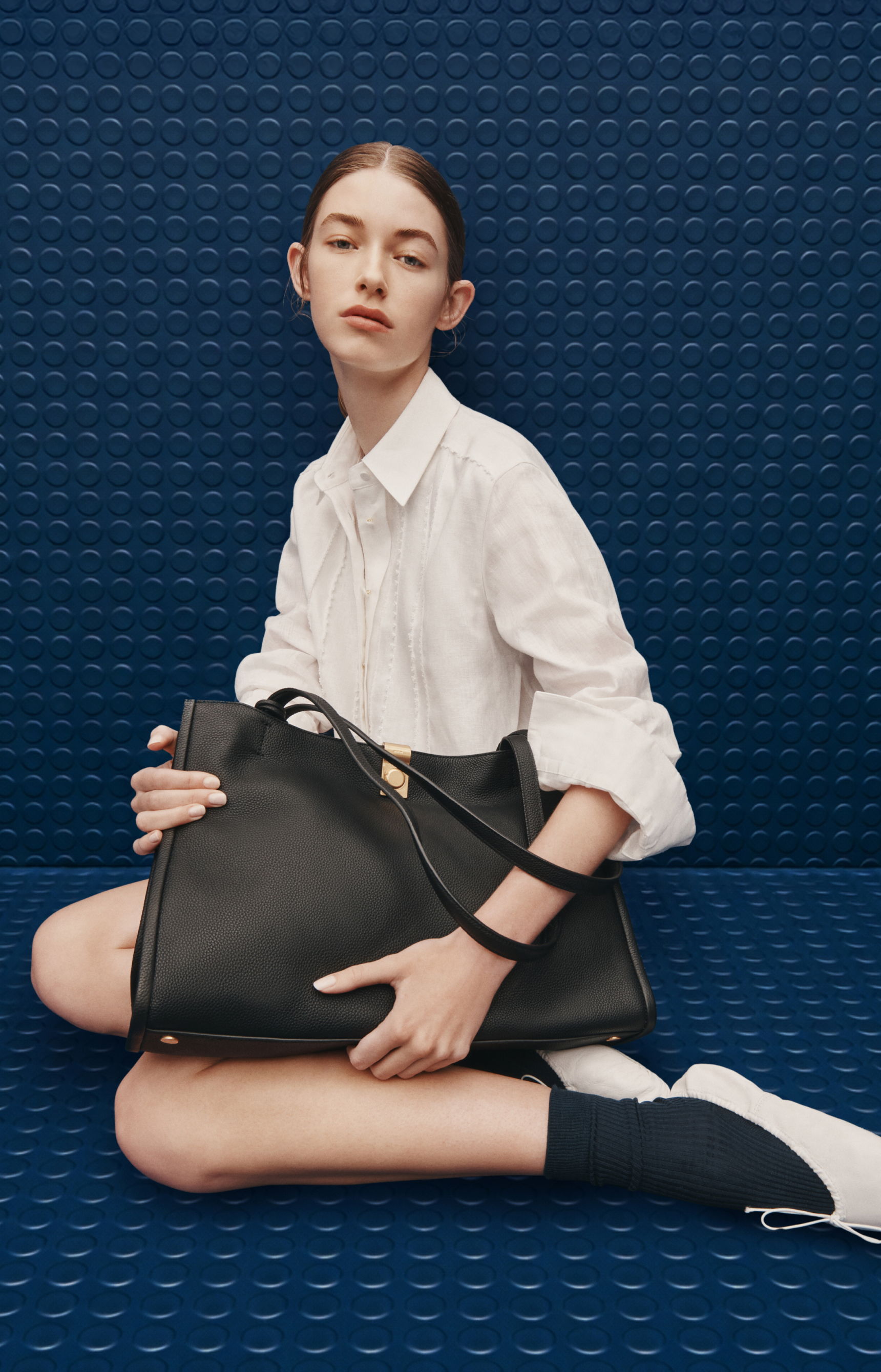 Model sitting with a black handbag against a blue background.