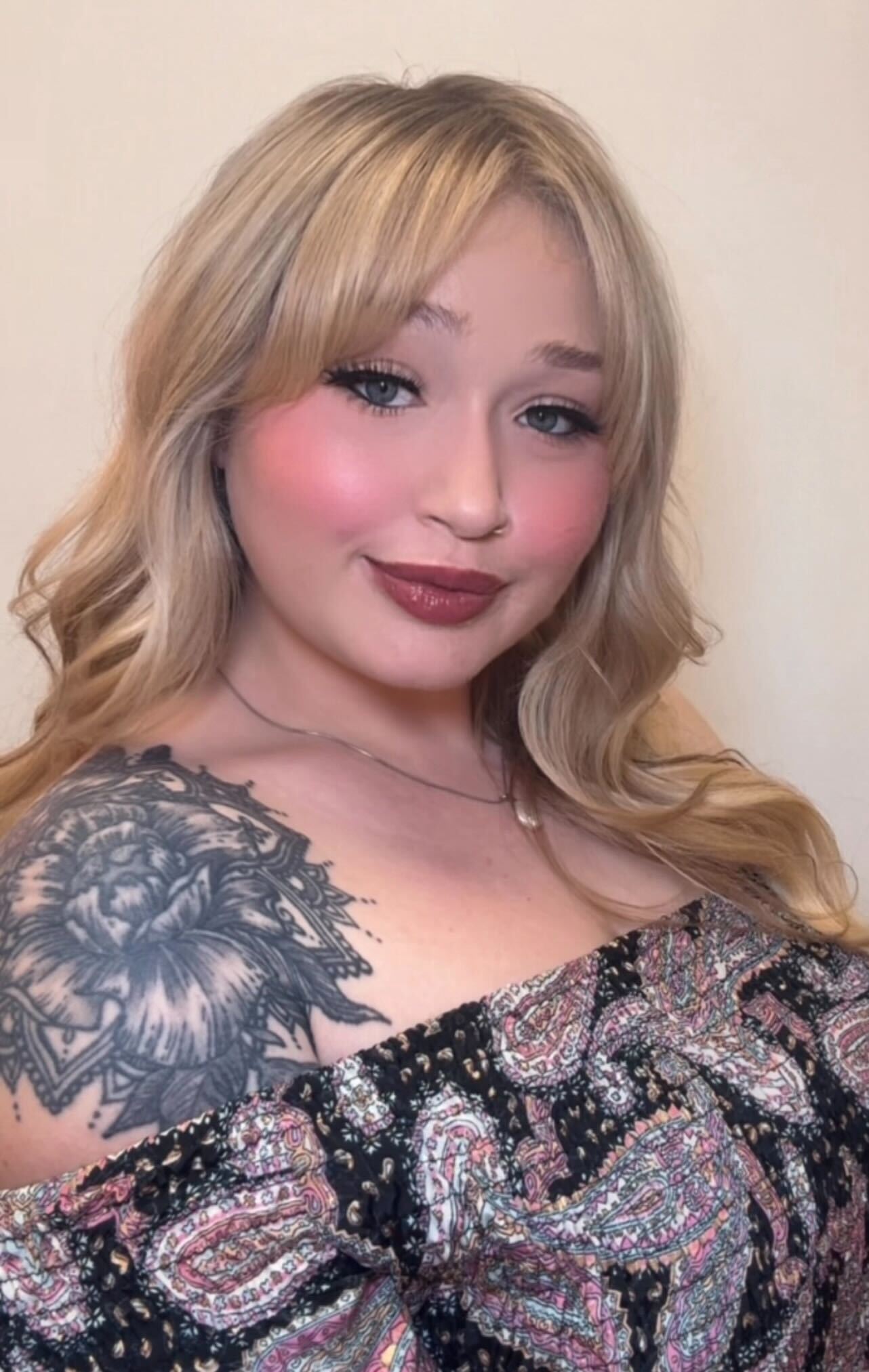 Young woman with blonde hair and floral tattoo, wearing a patterned top.