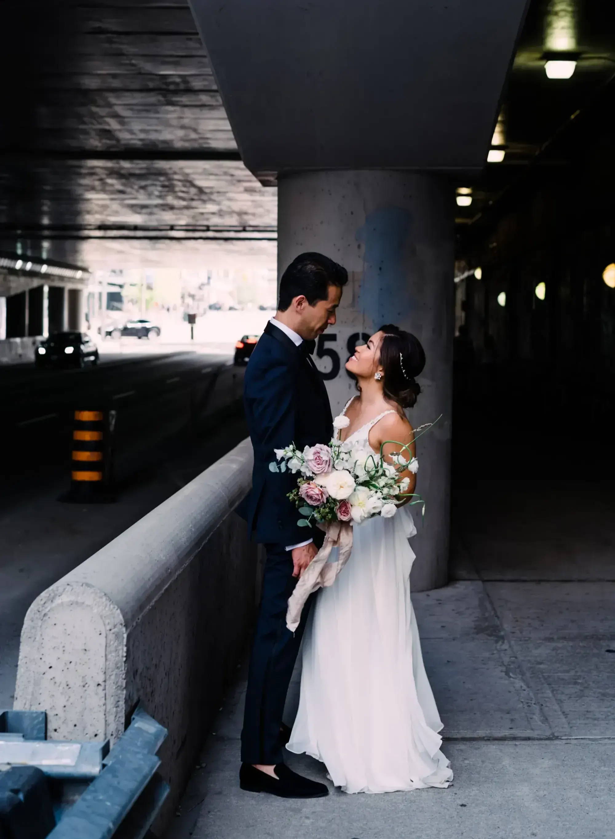 couple smiling at each other outside under an overpass, while bride holds a bouquet of blush and white florals