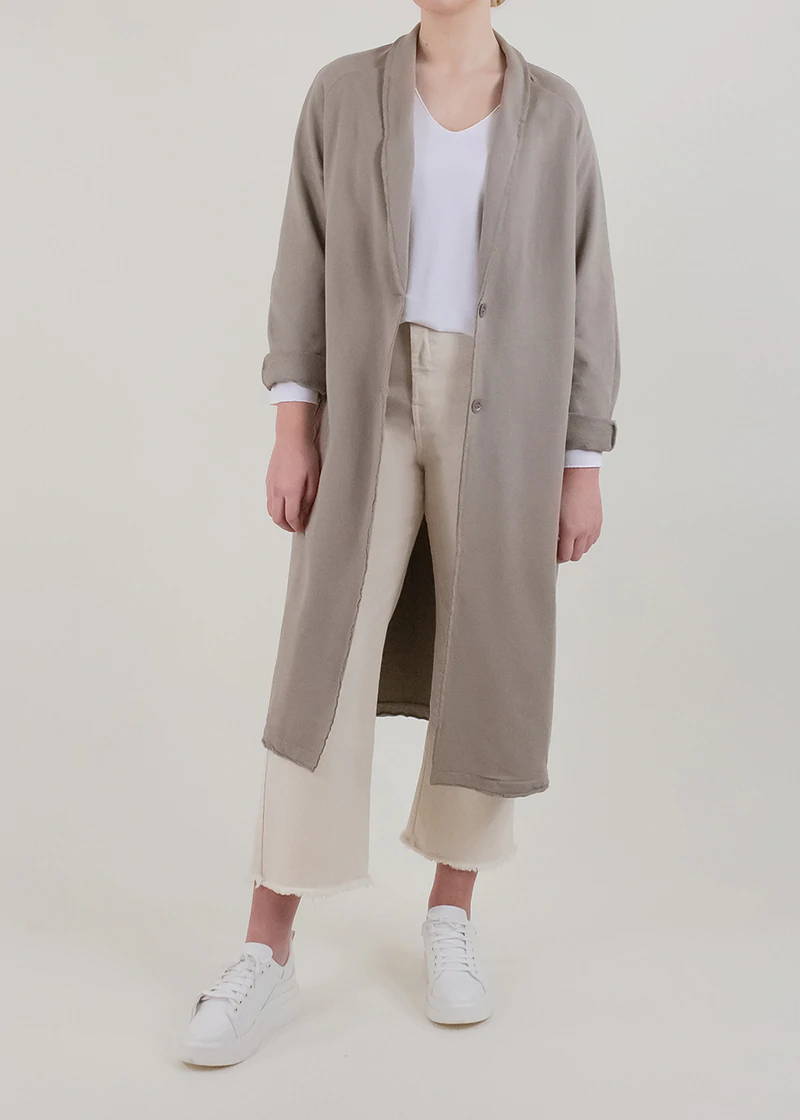 A model wearing a taupe trench coat over a white  jumper, cream trousers and white trainers