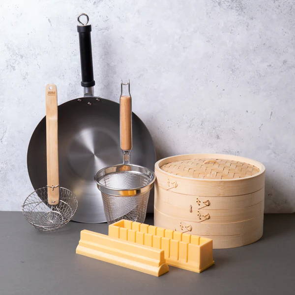 An oriental cooking set on the kitchen table. It includes a bamboo steamer, a wok and more.