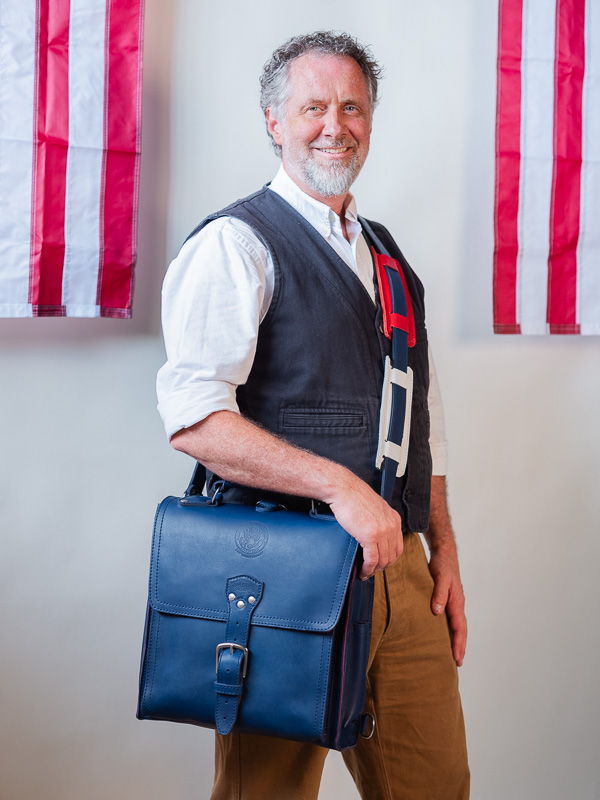 Man in a vest holding a blue bag, with flags in the background.