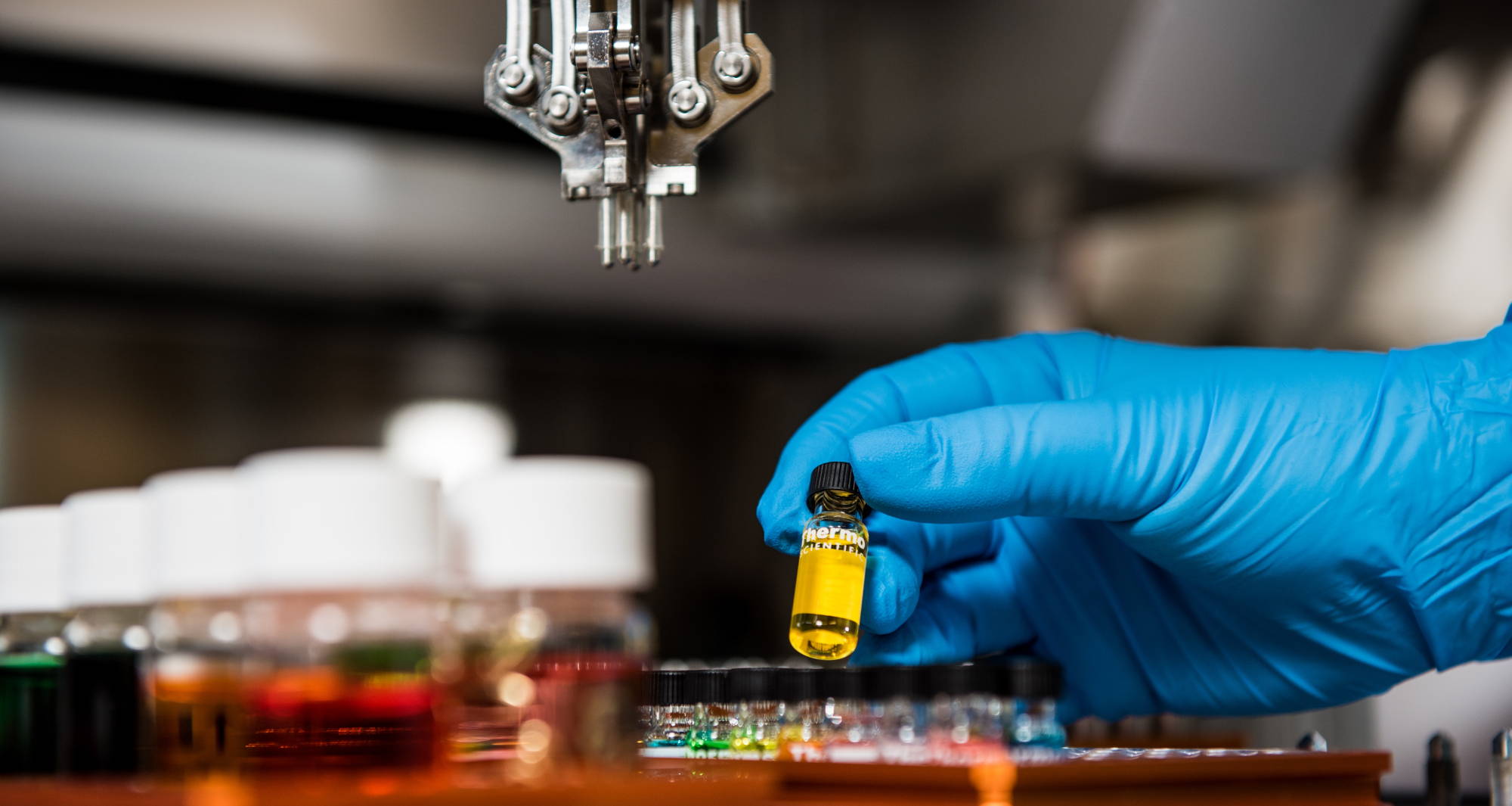 A hand in a blue rubber glove holds a small vial in a science lab.