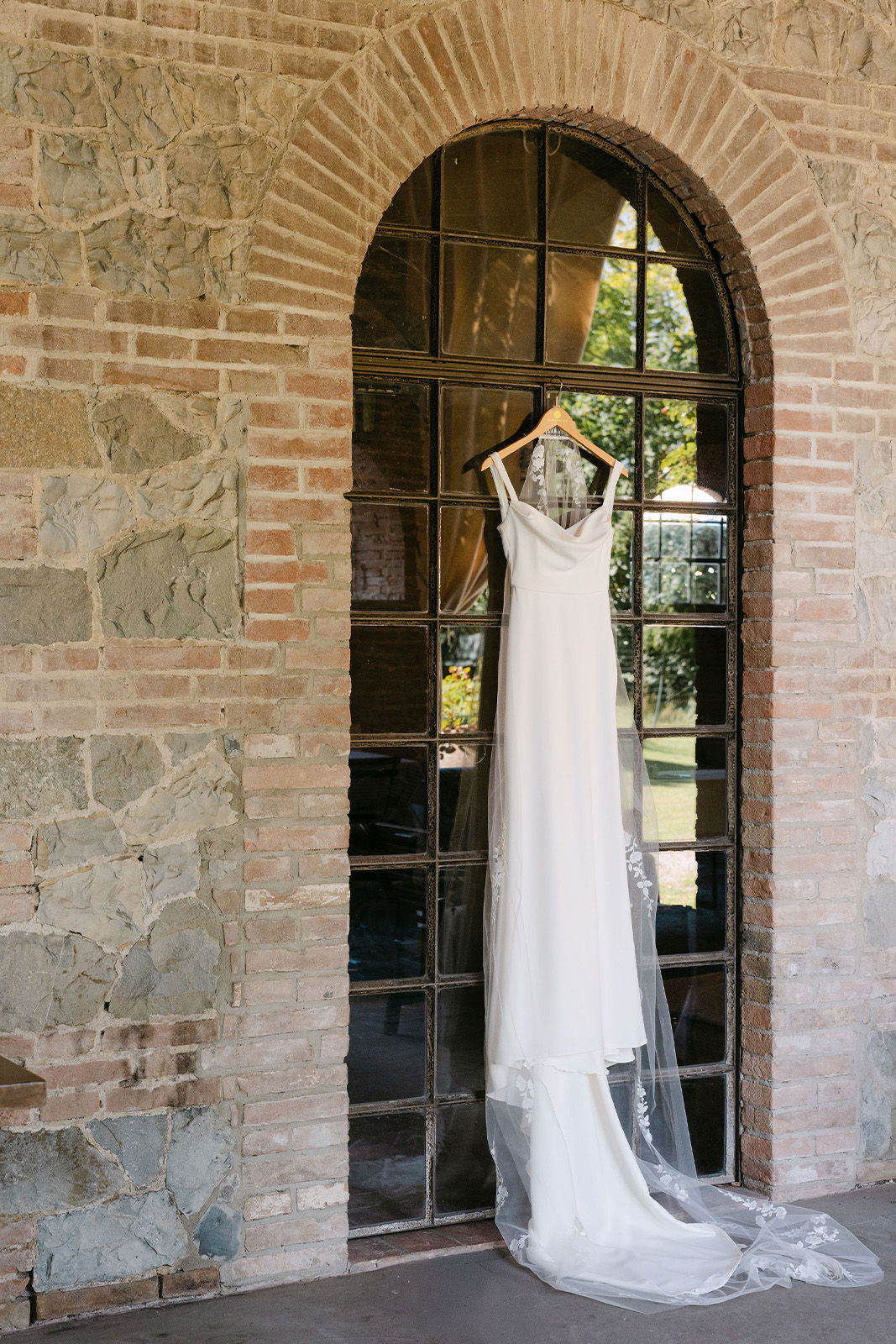 Wedding dress hanging in an arched window with a stone wall backdrop.