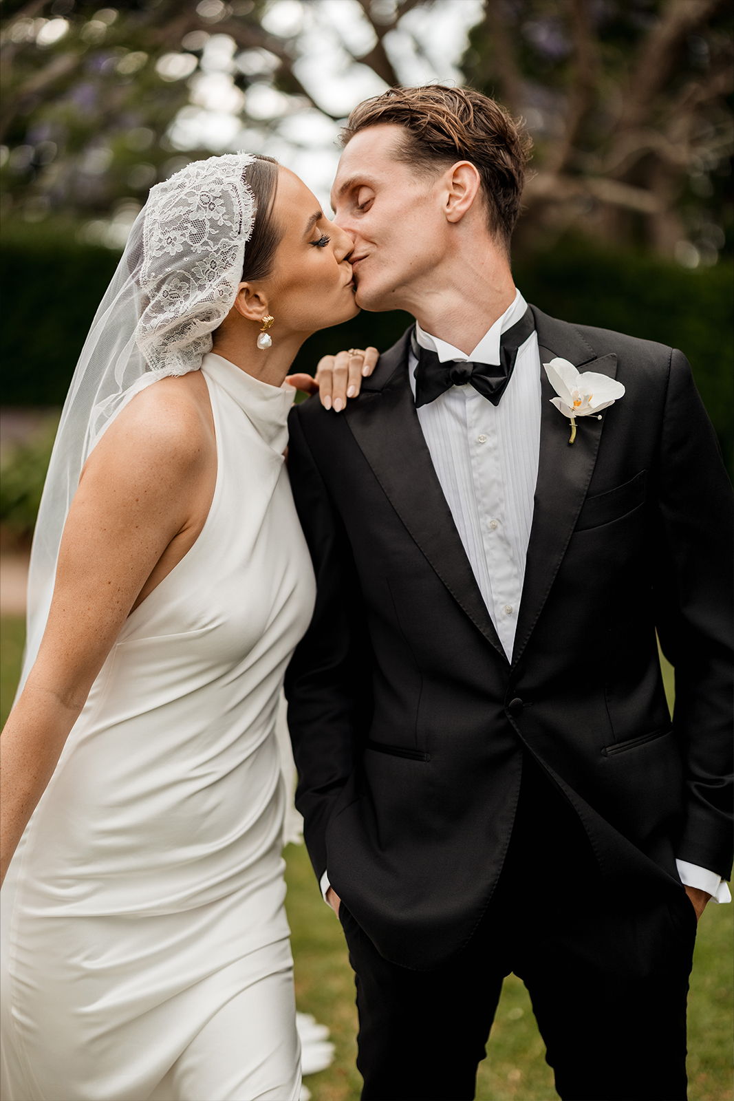 Bride and groom kissing in a garden setting, dressed in formal wedding attire.