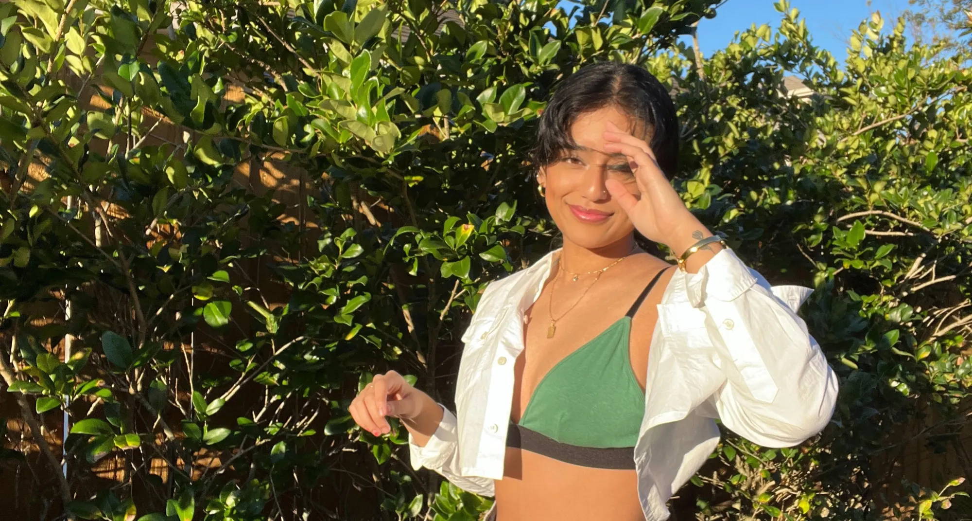 A woman wearing a green bralette, blue jeans, and an unbuttoned white shirt stands in front of green shrubbery. 