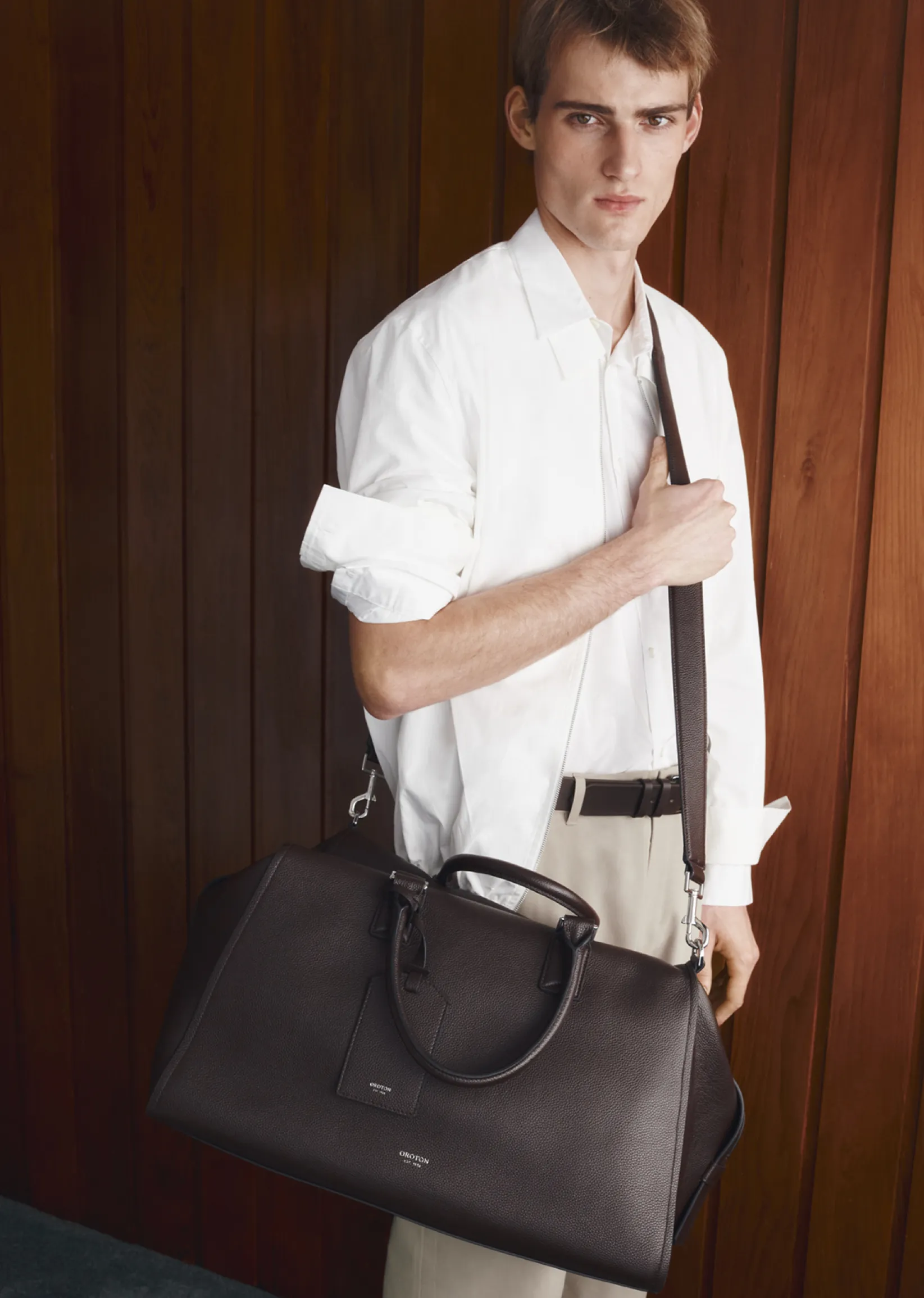 Young man holding a large black bag, wearing a white shirt and trousers.
