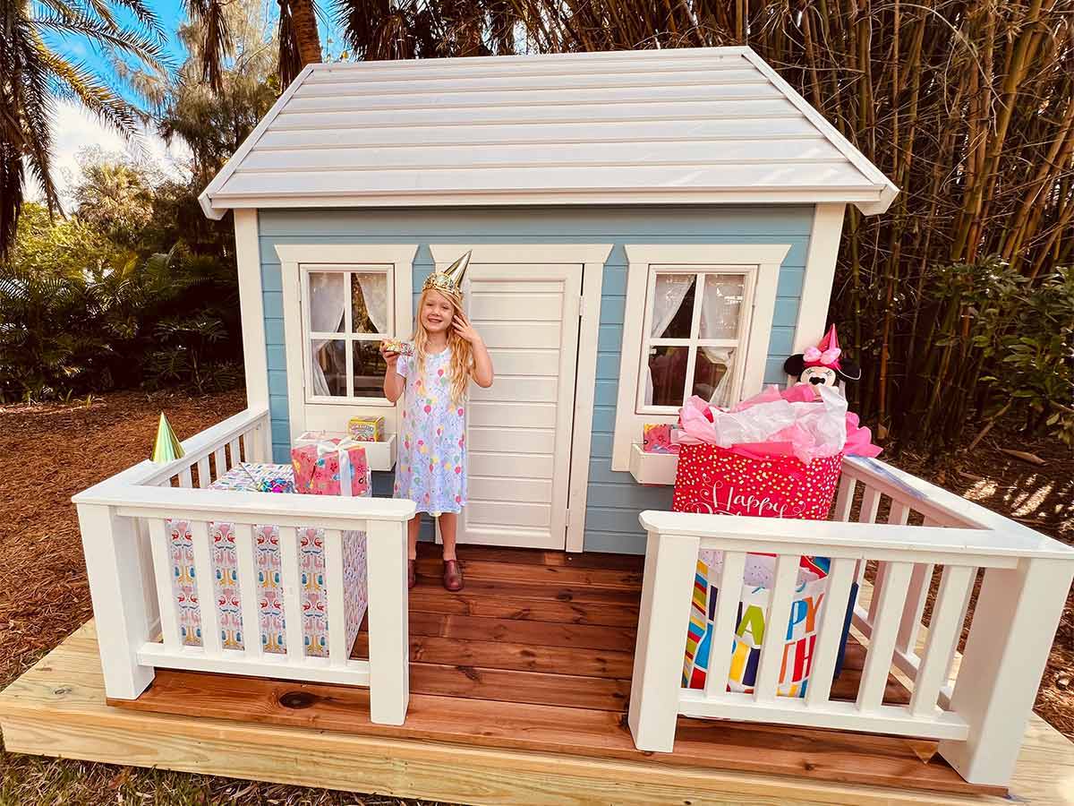 One girl standing on the terrace of a wooden playhouse with birthday presents by WholeWoodPlayhouses