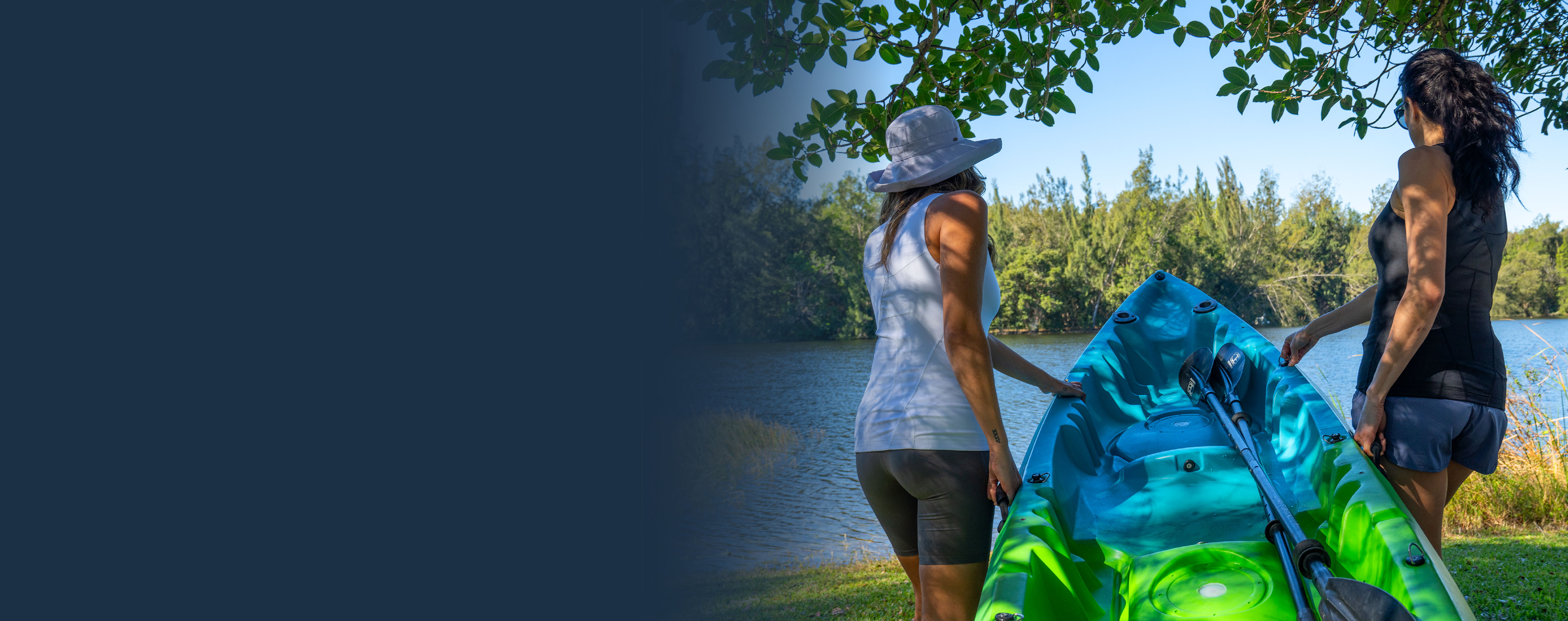 Two women wearing Tommie Copper compression while carrying a colorful kayak by a lake under trees.