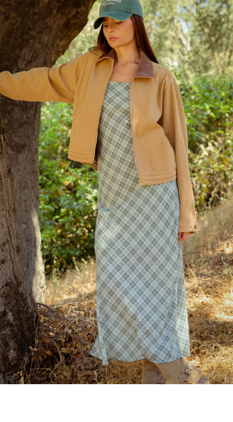 Woman wearing a long plaid dress and a beige jacket near a tree.