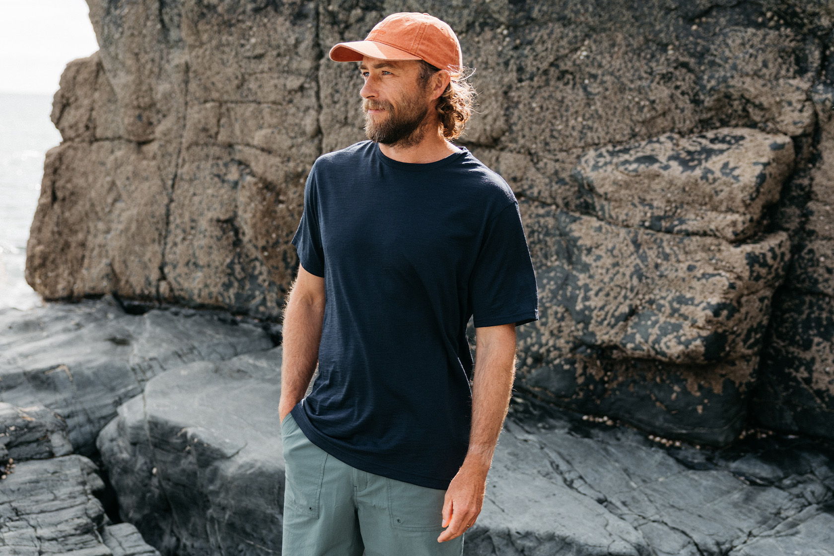Man standing on rocks near the ocean, wearing a hat and casual clothing.