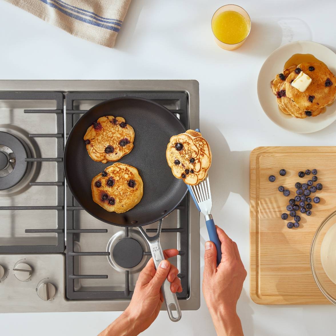 A bird’s eye view of a Misen Nonstick Pan on a stovetop with a chef’s hand flipping blueberry pancakes in it. A stack of pancakes sits on a plate to the right of the stovetop.