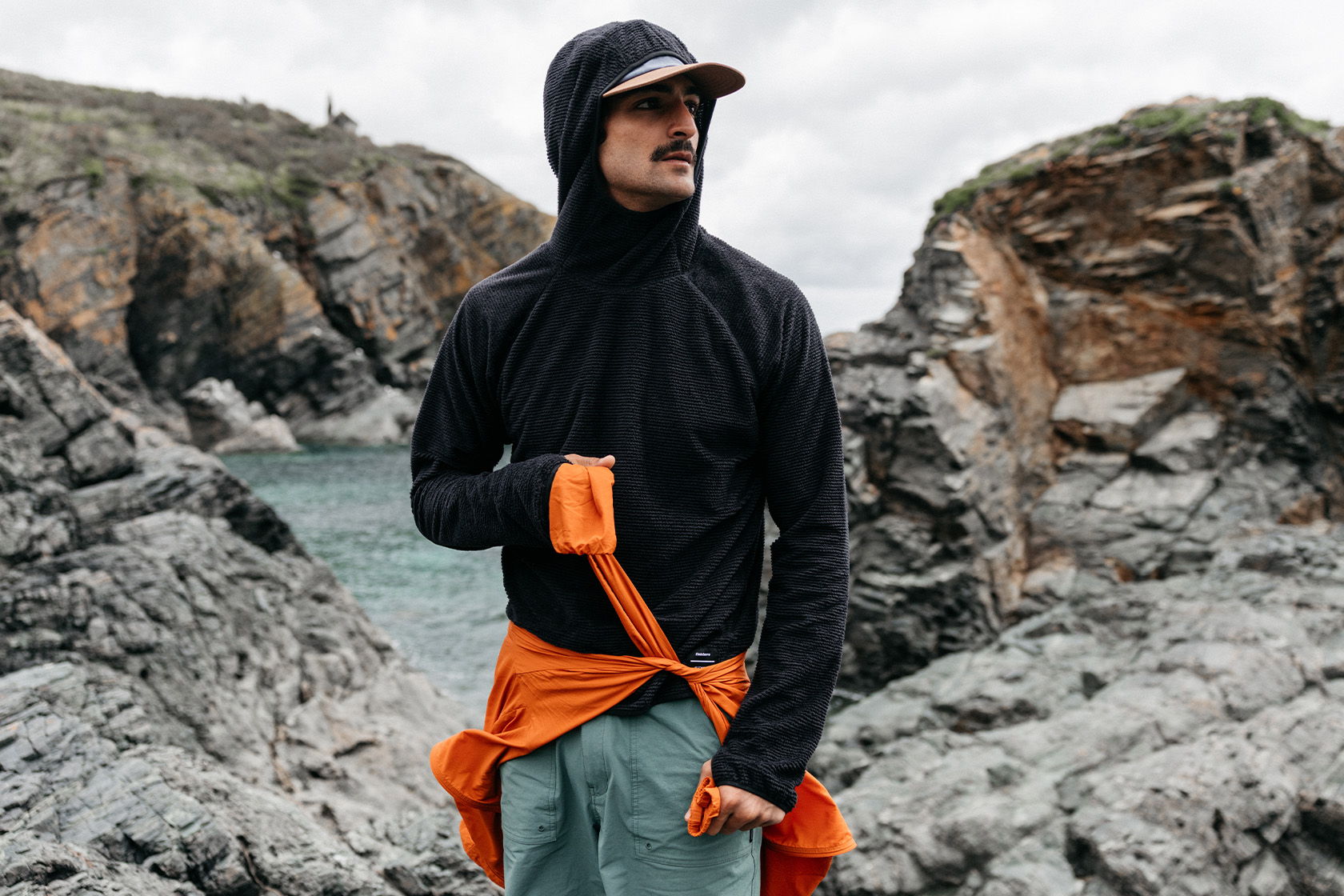 Person in a black hoodie with an orange shirt, standing on rocky terrain.