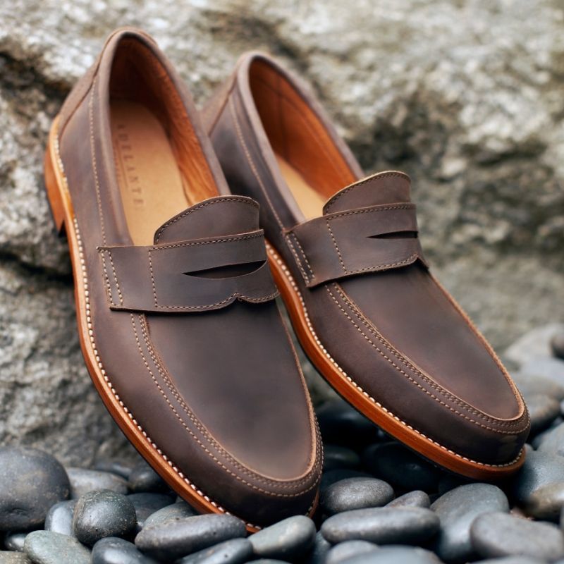 A pair of Adelante dark brown leather wide width penny loafers resting on smooth black river stones.