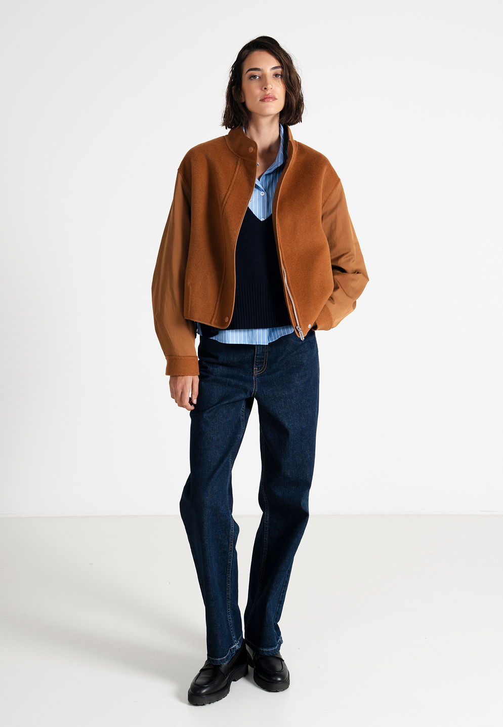 Person wearing a brown jacket, blue shirt, and dark jeans, standing against a white background.