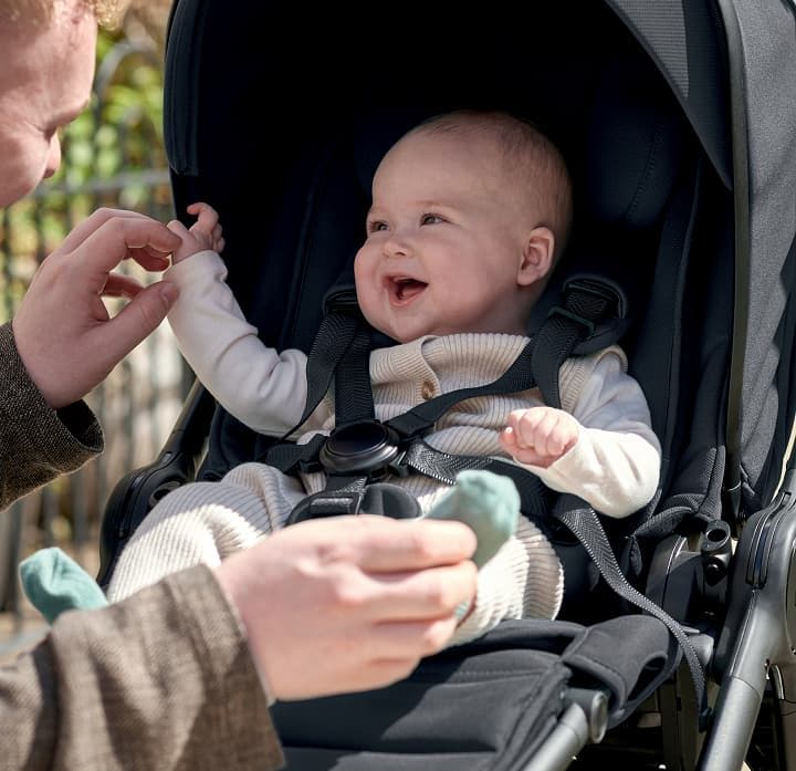 A smiling baby in a stroller reaches for an adult's hand.