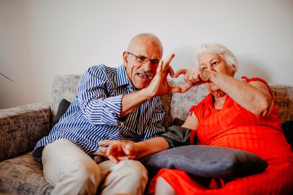 Elderly couple making heart shapes with their hands, sitting on a couch.