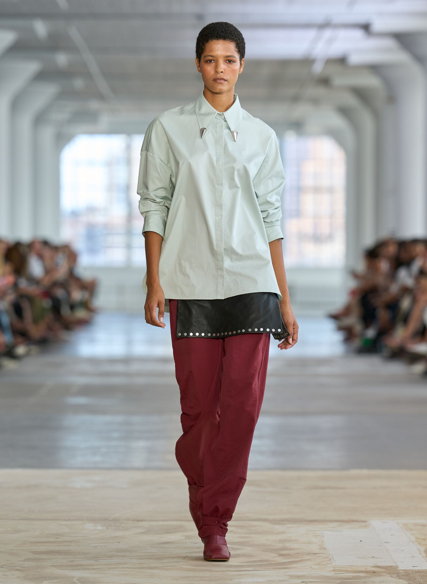 Model walking down a runway wearing a light shirt and burgundy pants.