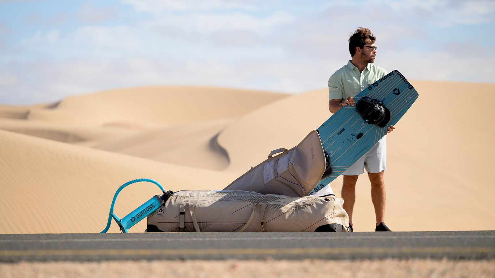 Person standing with a surfboard near sand dunes in a desert landscape.