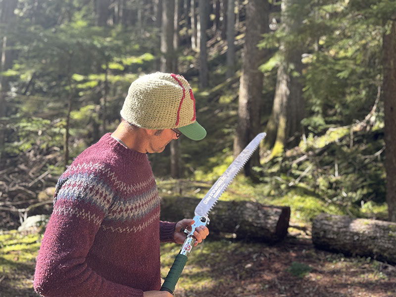 Person in a forest holding a large folding saw, wearing a hat and sweater.