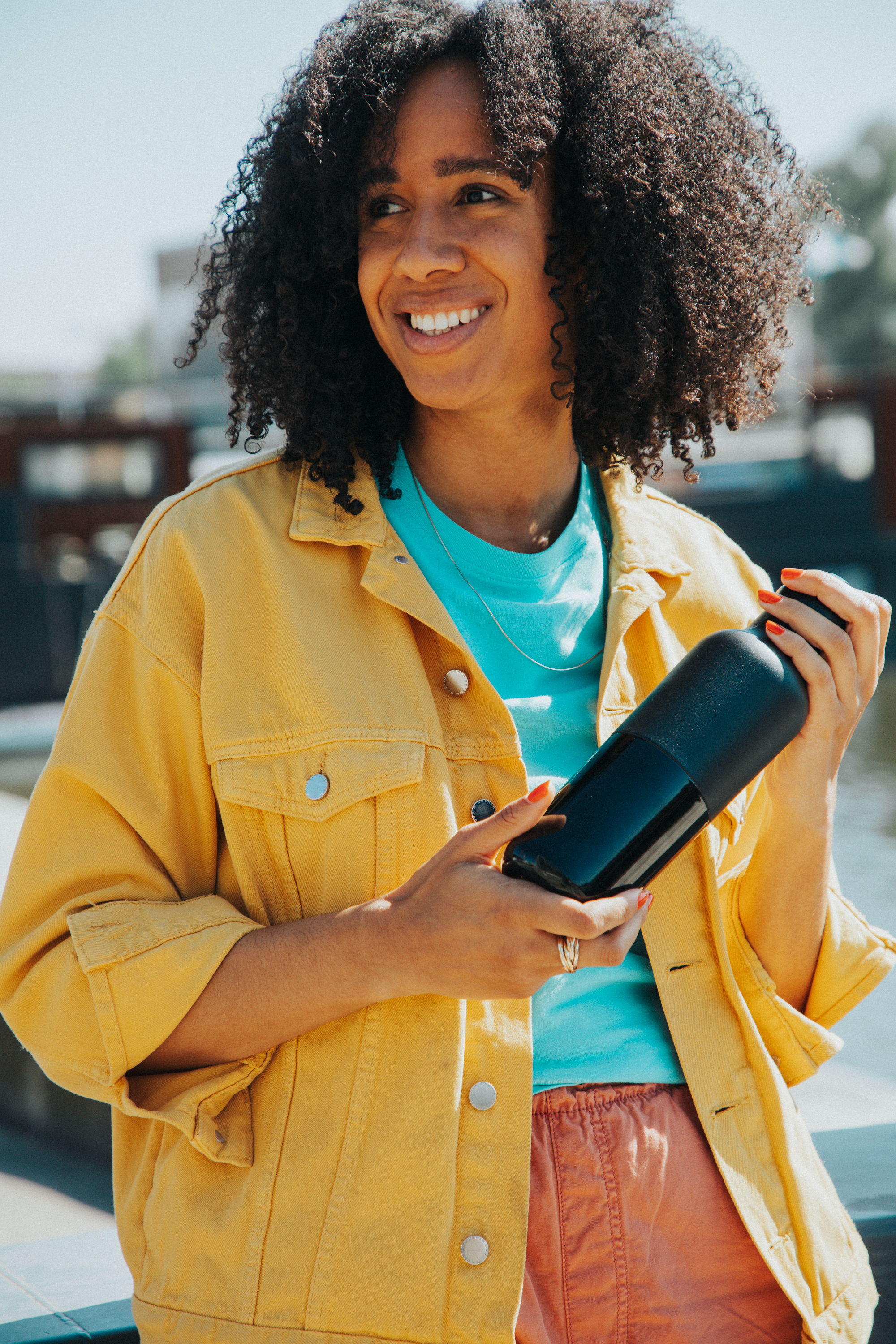 Smiling woman holding a black object, wearing a yellow jacket and colorful shirt.