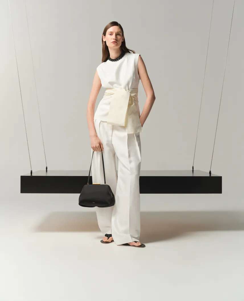 Model wearing white outfit, holding a black bag, against minimalist background.