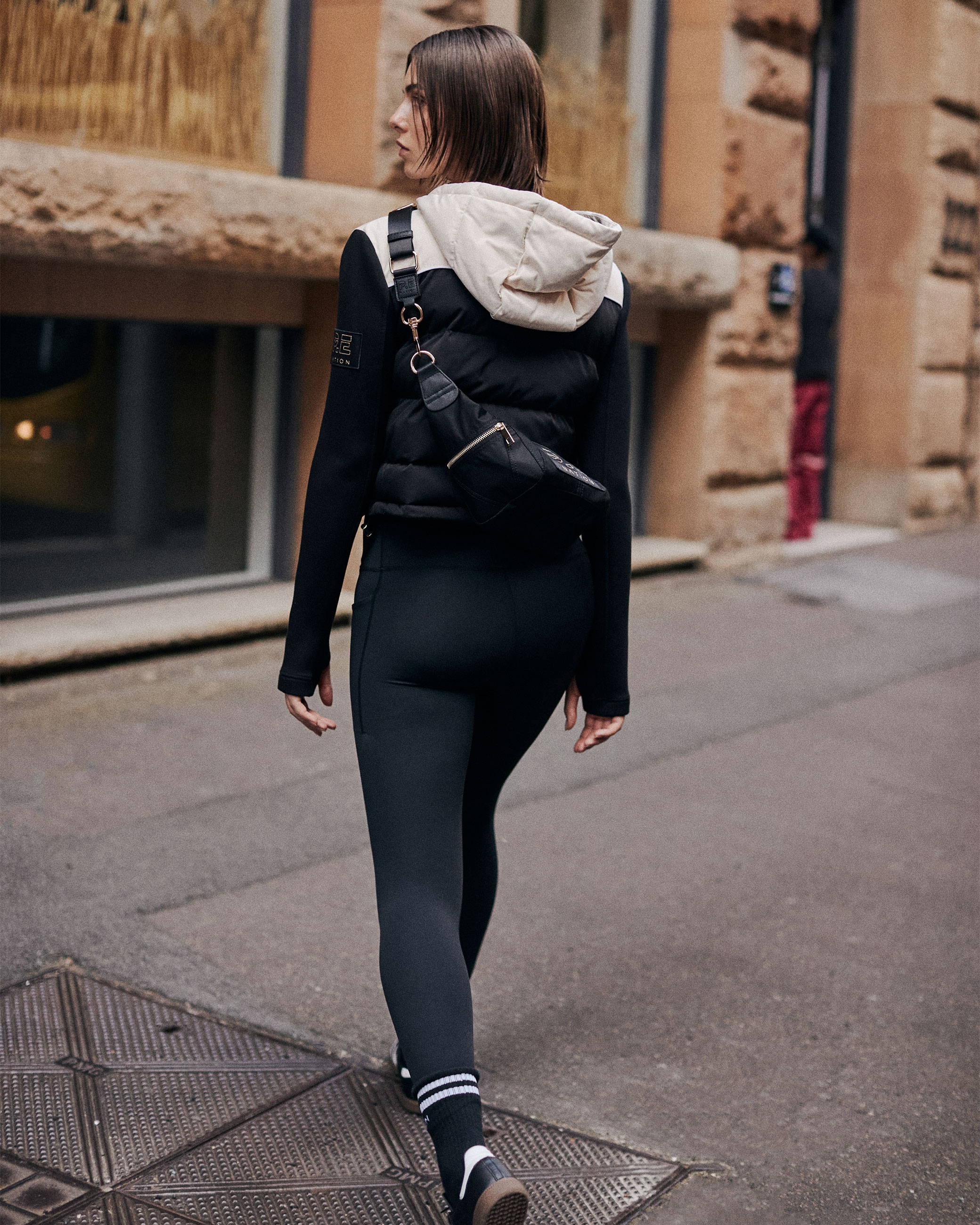 A girl is walking in the city with her back to the camera. She is wearing leggings, a jacket and a cross body bag.